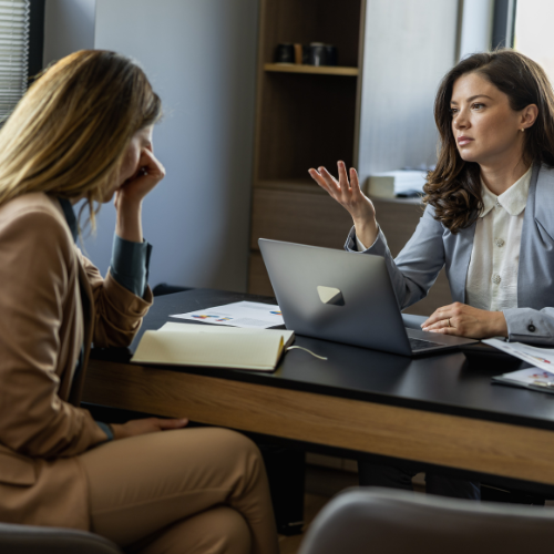 Two women talk to each other, the one on the left is frustrated and the woman on the right is wearing grey and has a laptop.